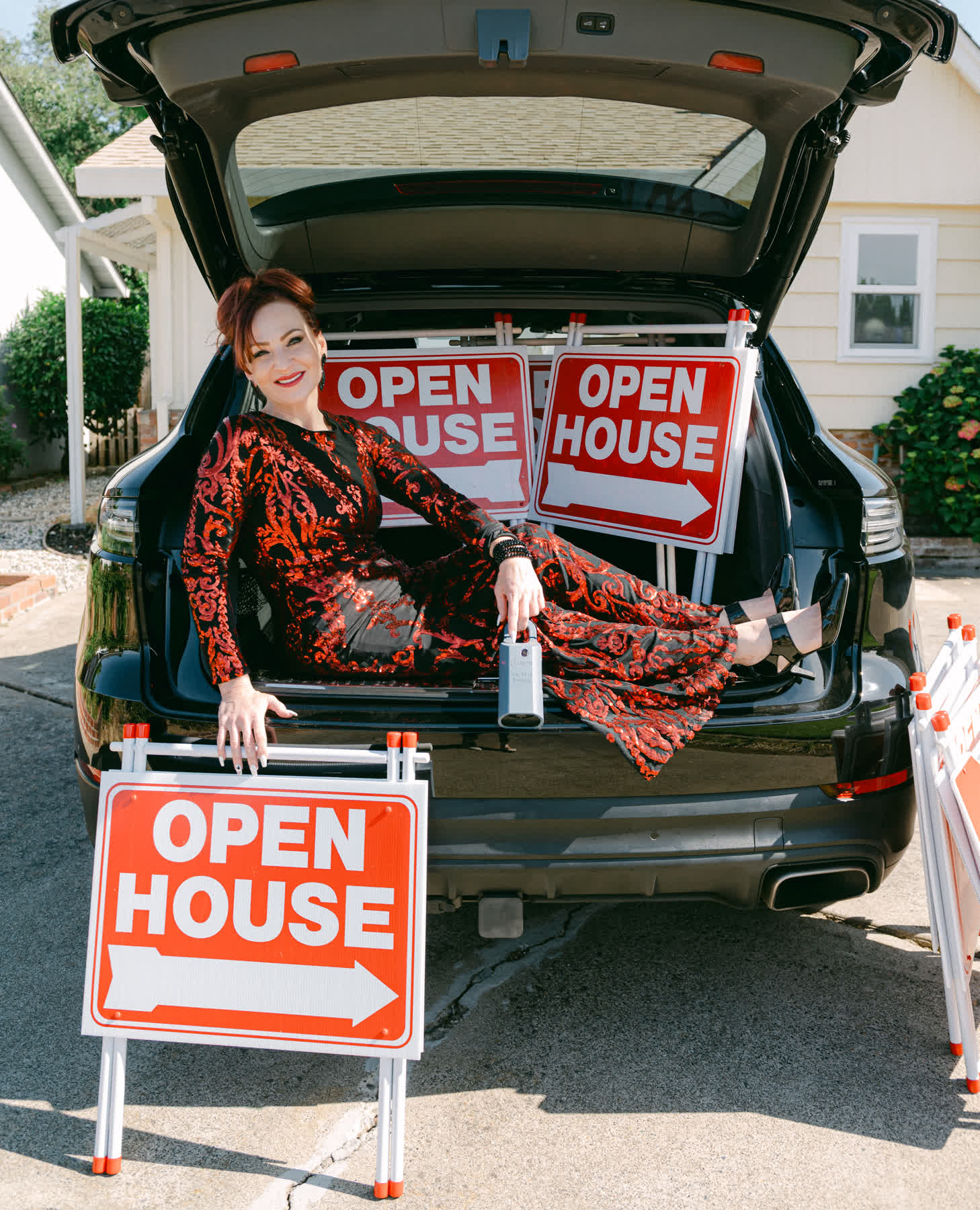 Dana Miller in a red sequined gown, reclining in the open trunk of her car beside three Open House signs
